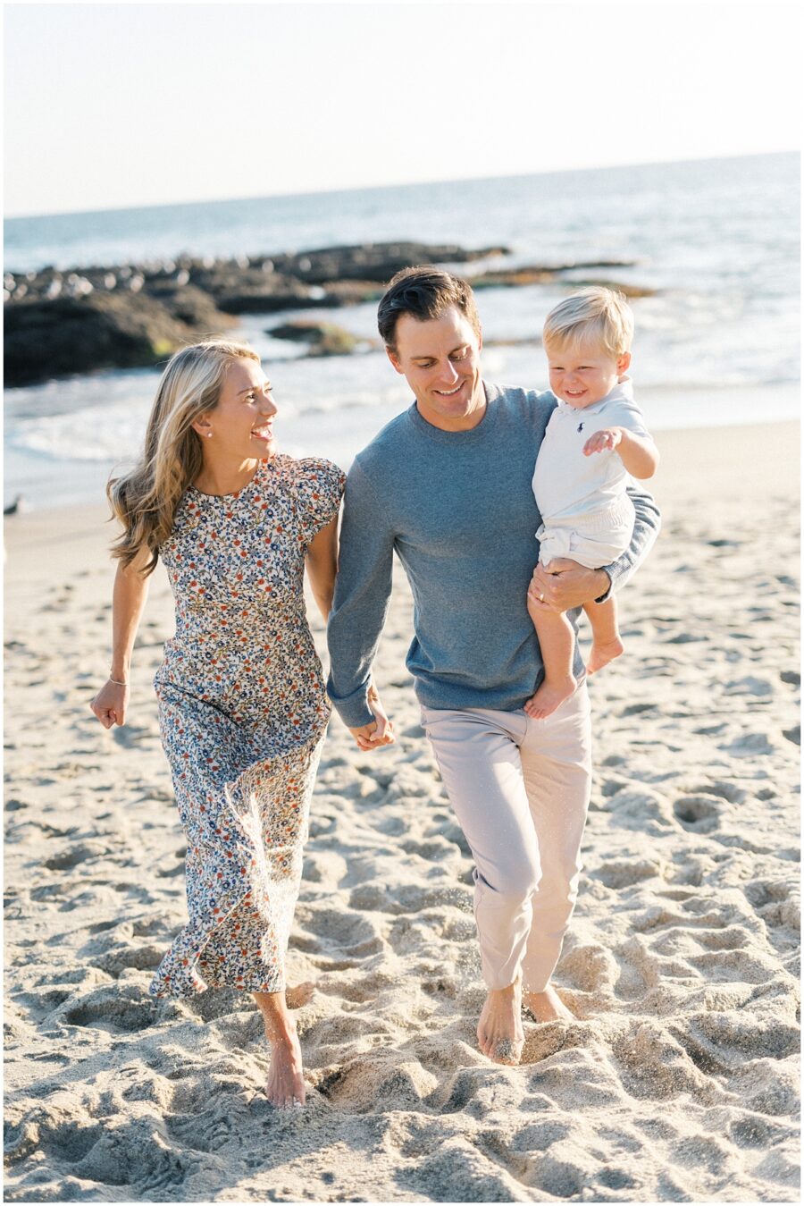 Mom wearing a floral dress and dad with toddler running on the beach in Laguna with toddler laughing.