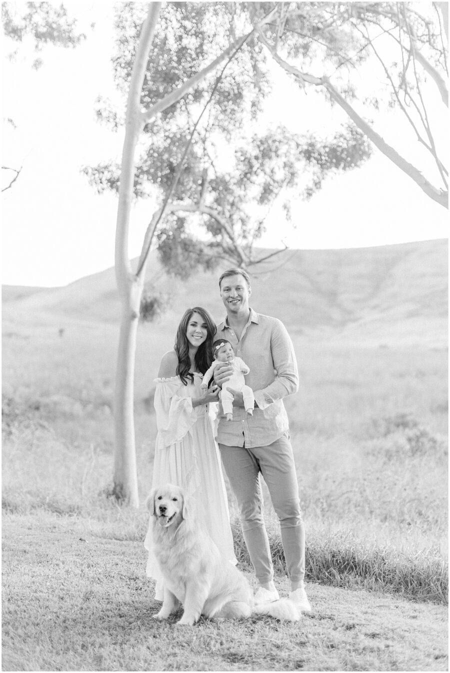 mom in long dress, dad holding newborn in grassy field with golden retriever