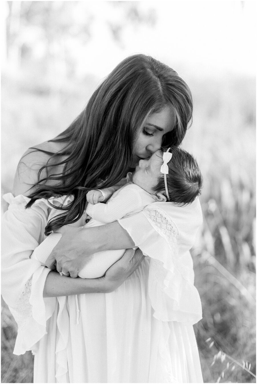 mom in white dress kissing newborn baby outdoors in grassy field in Irvine