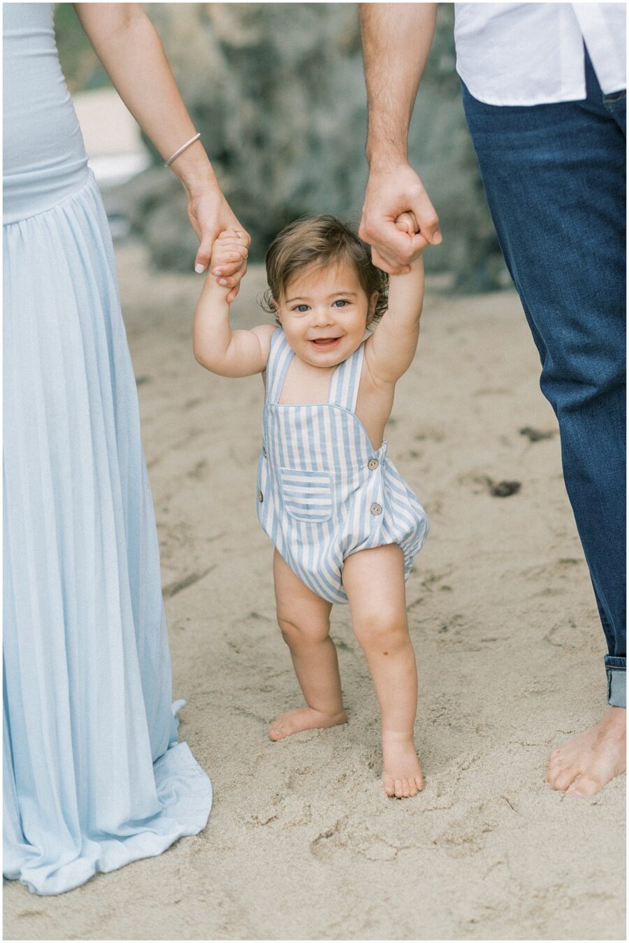 Baby boy in blue striped jumper in Laguna Beach holding parents hands on the sand.