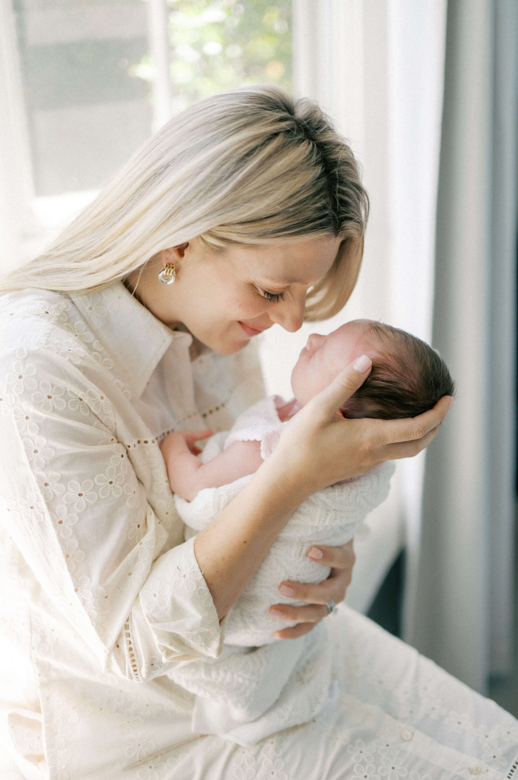 A happy mother in a white dress holds her newborn up to her face for nose nuzzles after visiting baby boutiques in newport beach while sitting in a window
