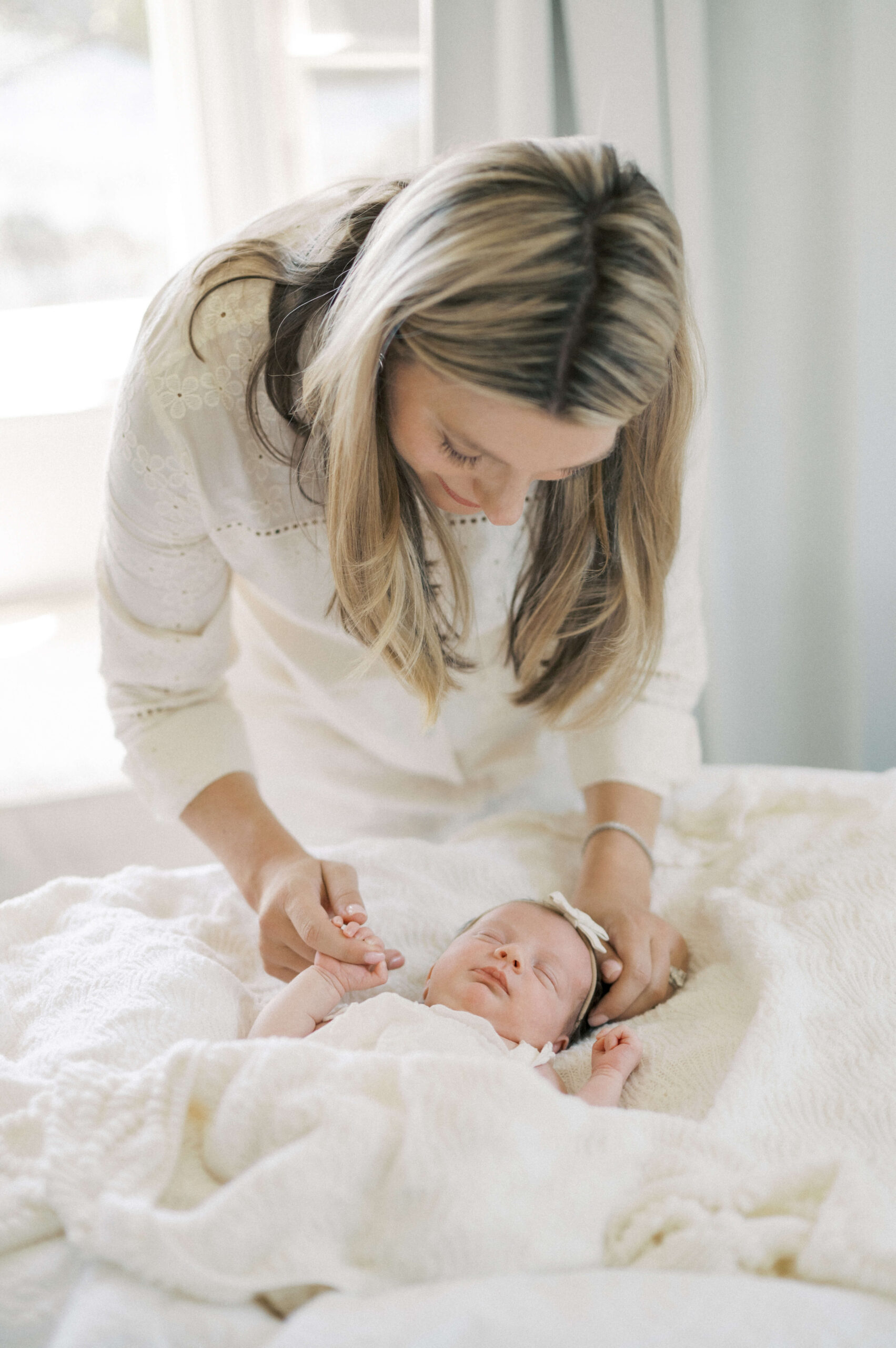 A mother in white leans down to hold her sleeping newborn's hand on a bed under white blankets after visiting baby boutiques in newport beach