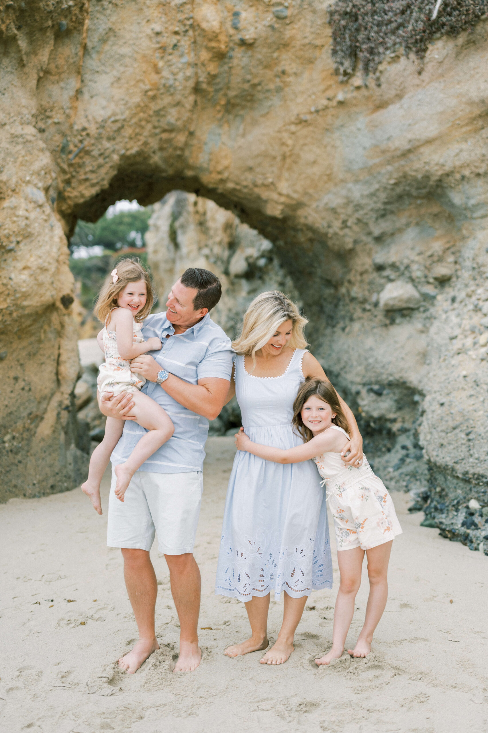 A mom and dad in blue hug and lift their toddler daughters on a beach in front of a natural arch