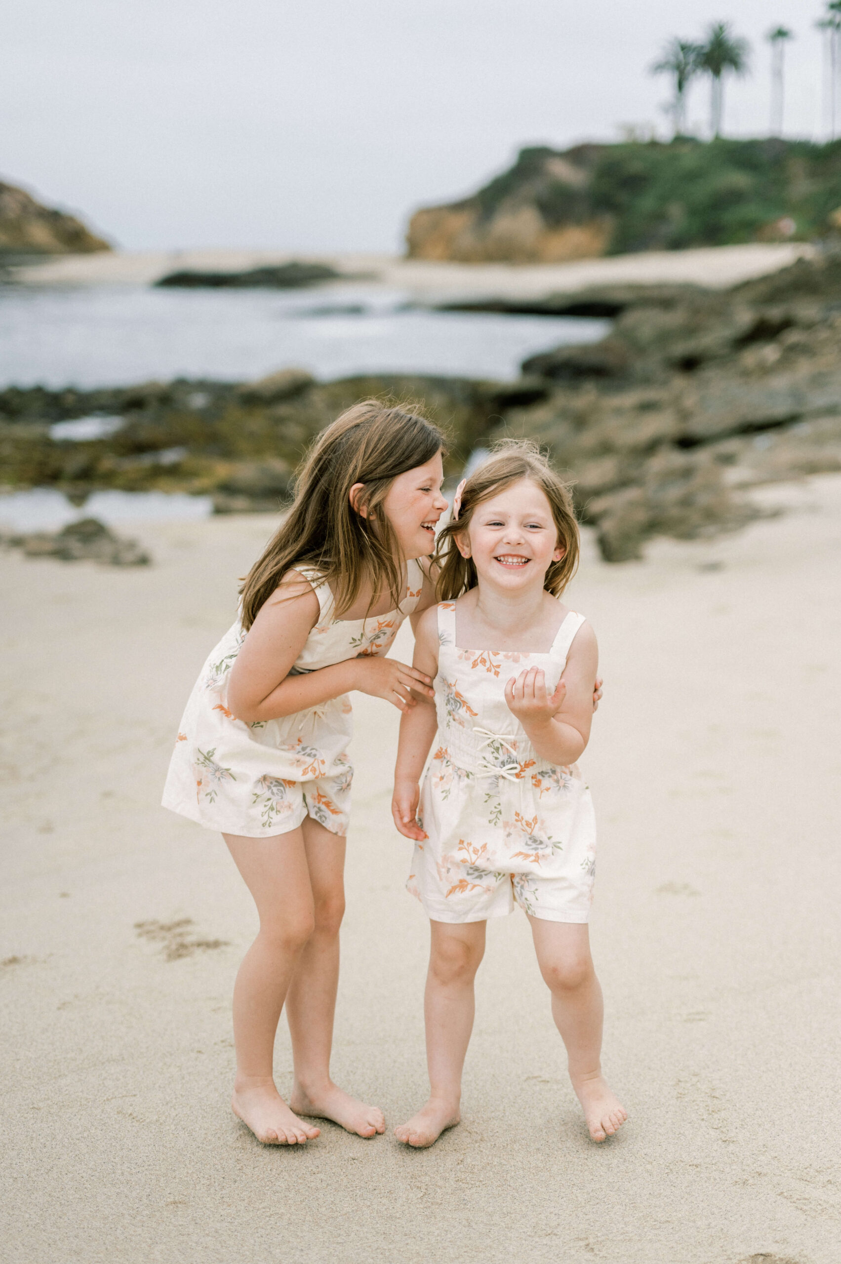 Toddler sisters in matching floral print overalls laugh and play on a beach with big smiles