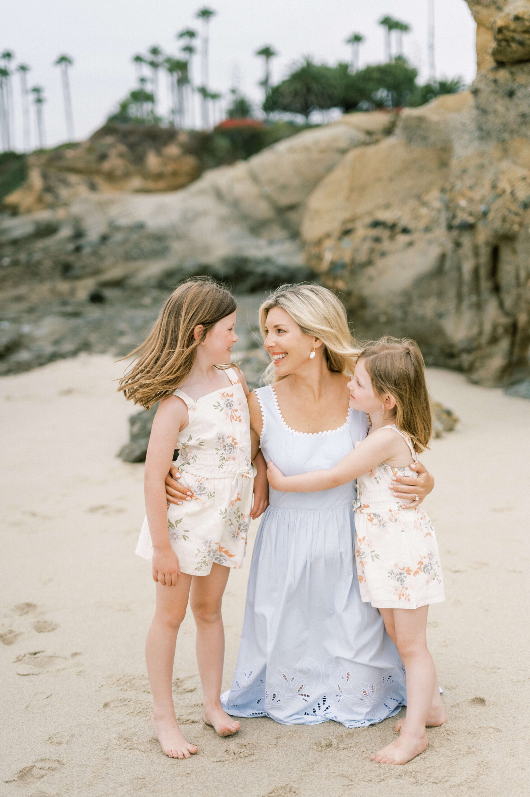 A smiling mother in a blue dress hugs her two toddler daughter in matching floral print outfits on a beach before visiting kid friendly restaurants in newport beach