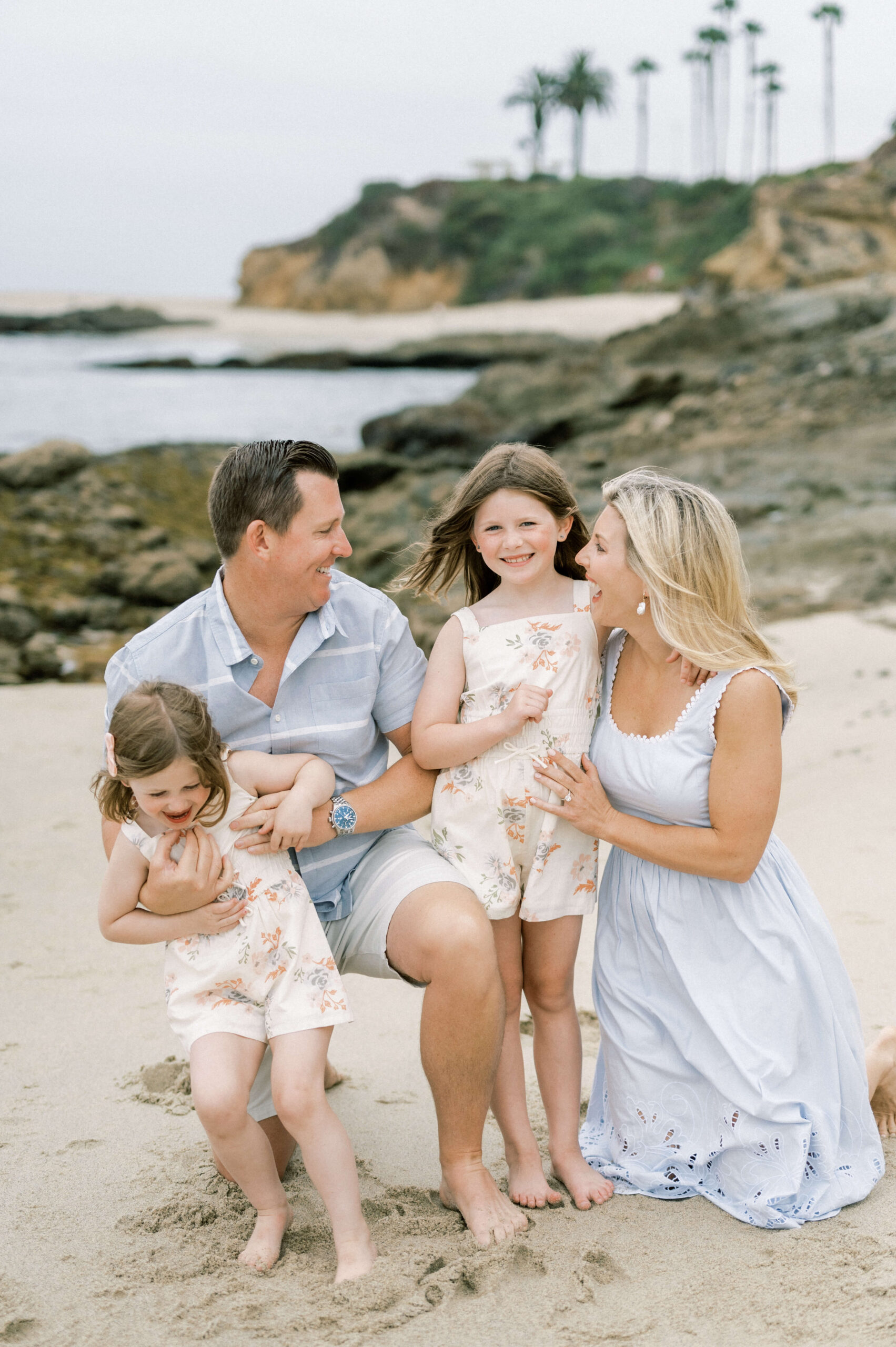 A happy mom and dad in blue outfits laugh and play with their two toddler daughters on a beach in matching floral jumpers before visiting kid friendly restaurants in newport beach