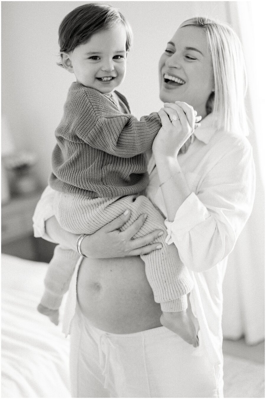 mom who is pregnant holding her toddler with his legs wrapped around her bare belly and both are smiling in the home.