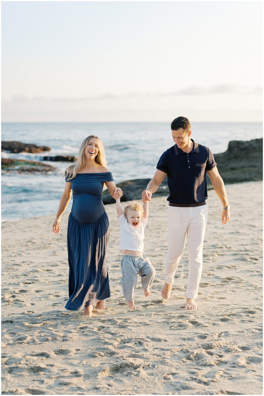 Pregnant mom and husband swinging their toddler on the beach at The Montage Laguna Beach laughing and smiling