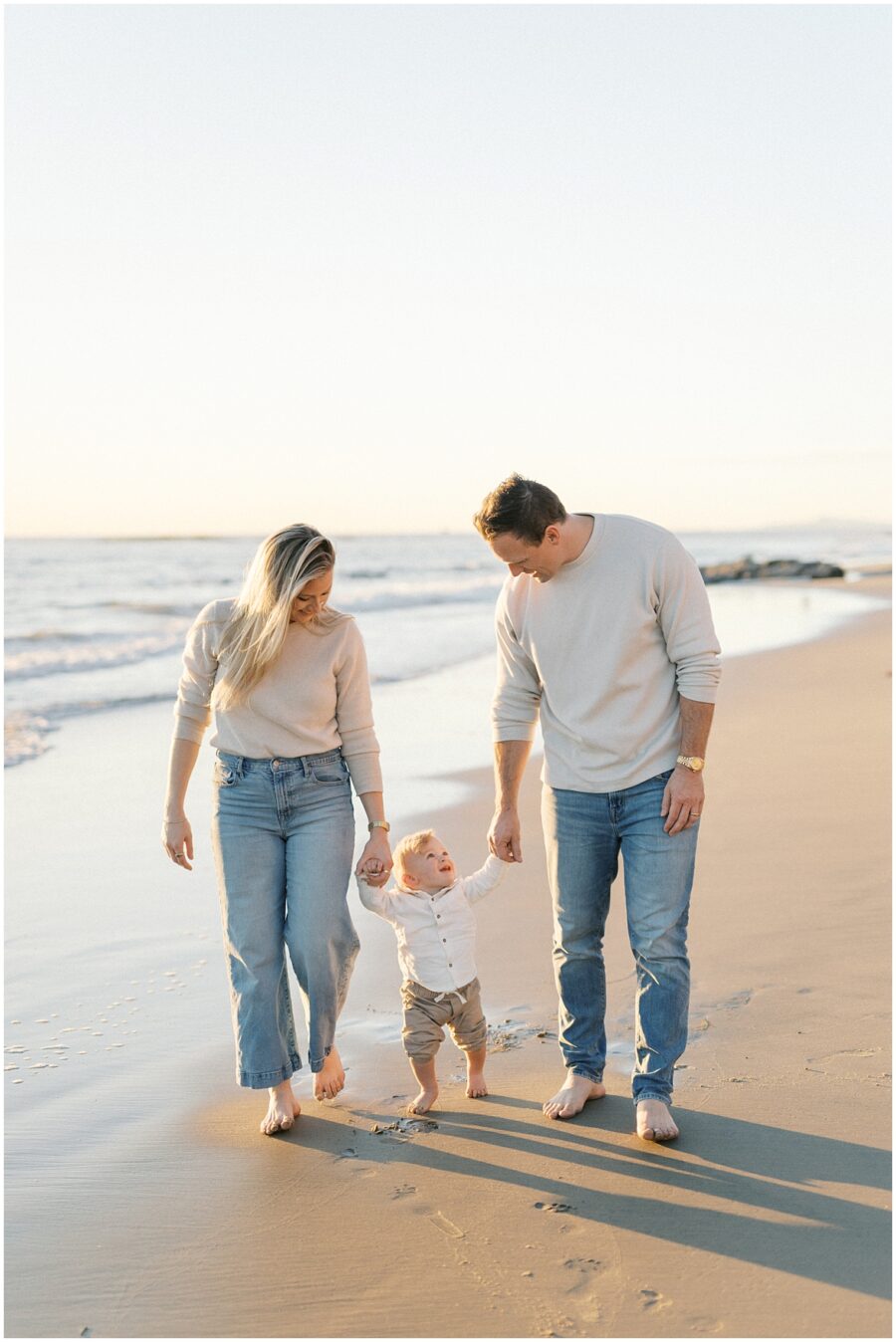 Family with toddler walking on the sand in Orange County with toddler taking his first steps on the beach.