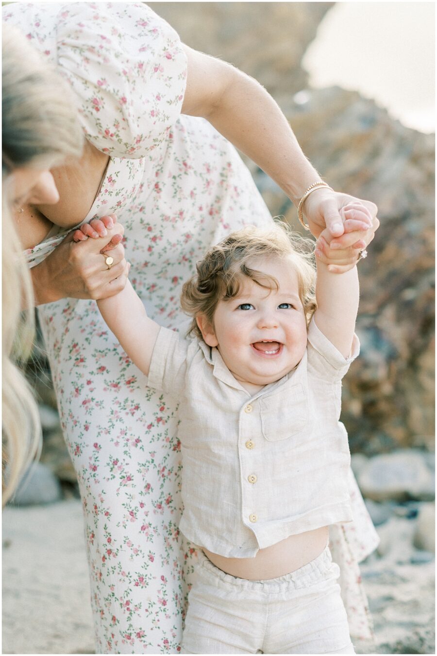 Mom holding toddlers in linen button down shirt hands while he smiles in Newport Beach